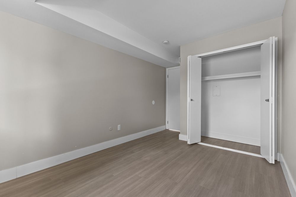 Bright bedroom with hardwood floors, neutral gray walls, white baseboards, and an open bifold closet door revealing built-in shelving.