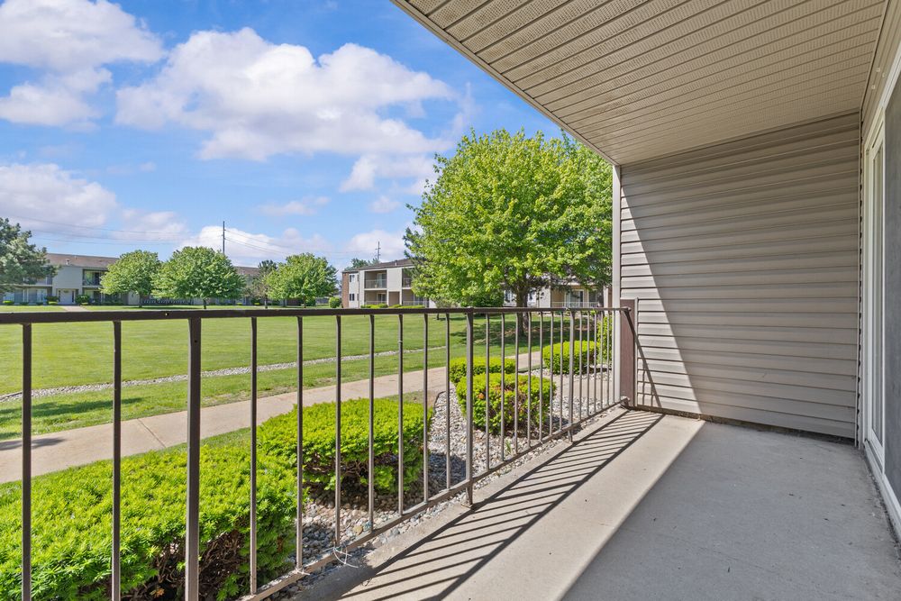 Sunny apartment balcony with metal railing overlooking lush green lawn and trees under a blue sky.