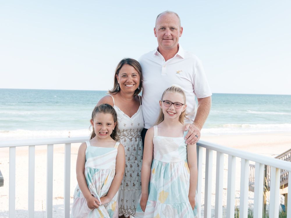 Family of four smiling together on a beach boardwalk with ocean in the background.