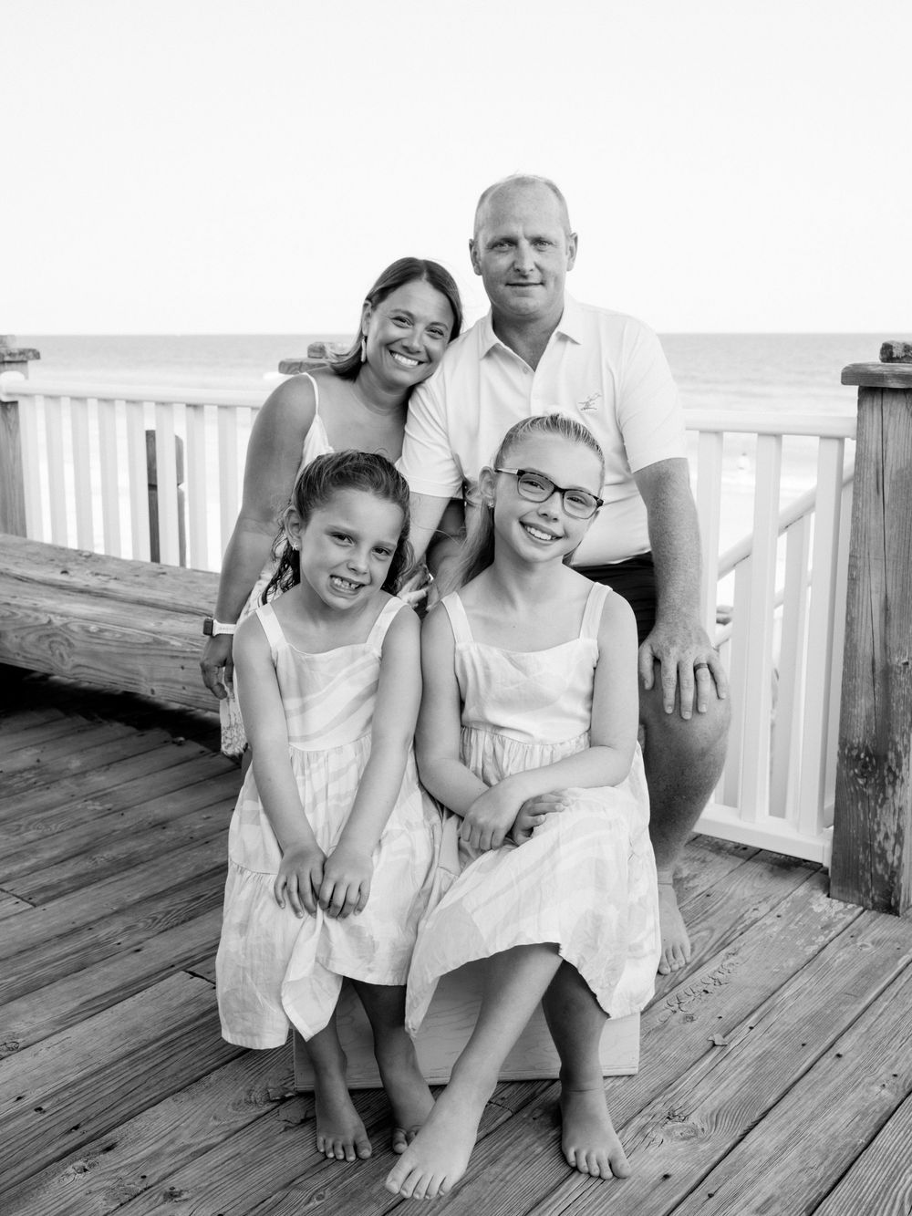 Black and white family portrait of two adults and two girls in dresses posing on a beach boardwalk.