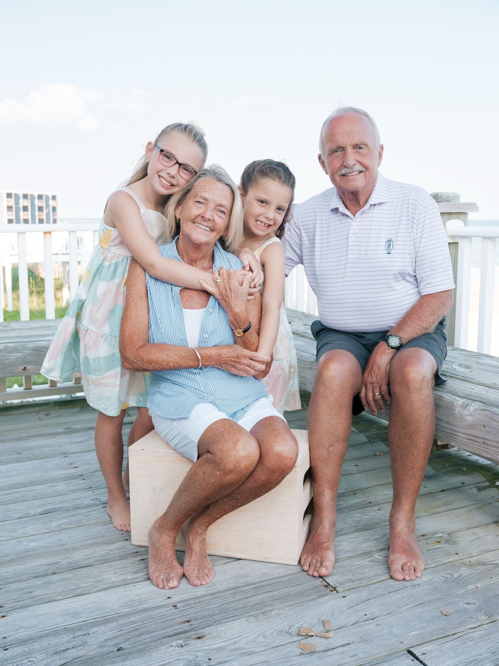 Grandparents sit on wooden deck with two young granddaughters hugging them at the beach.