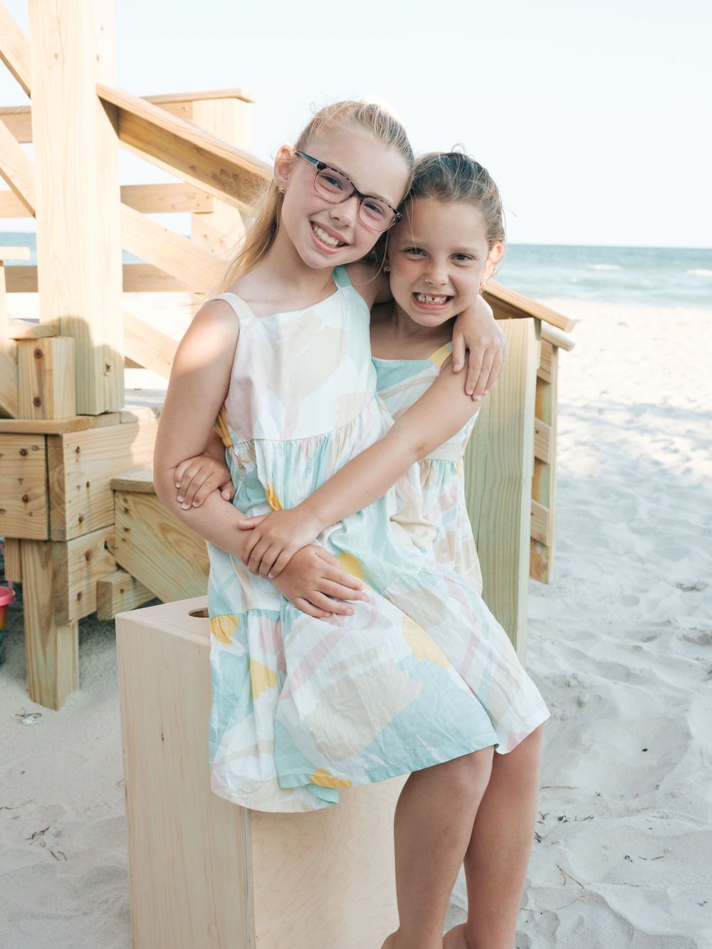 Two young sisters in matching pastel dresses embrace on a sunny beach boardwalk.
