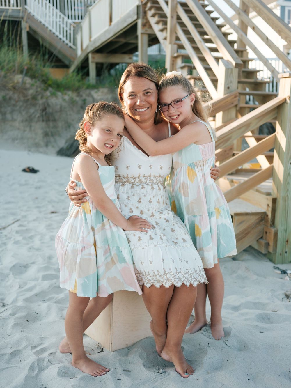 Woman in white dress hugging two girls in matching pastel dresses on a sandy beach.