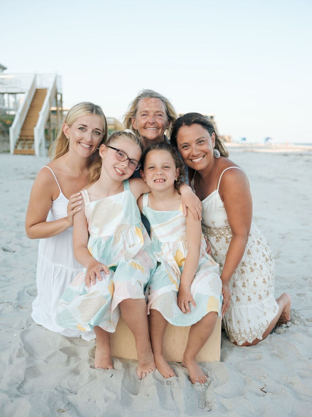 Group of five females posing together on beach in white and pastel outfits.