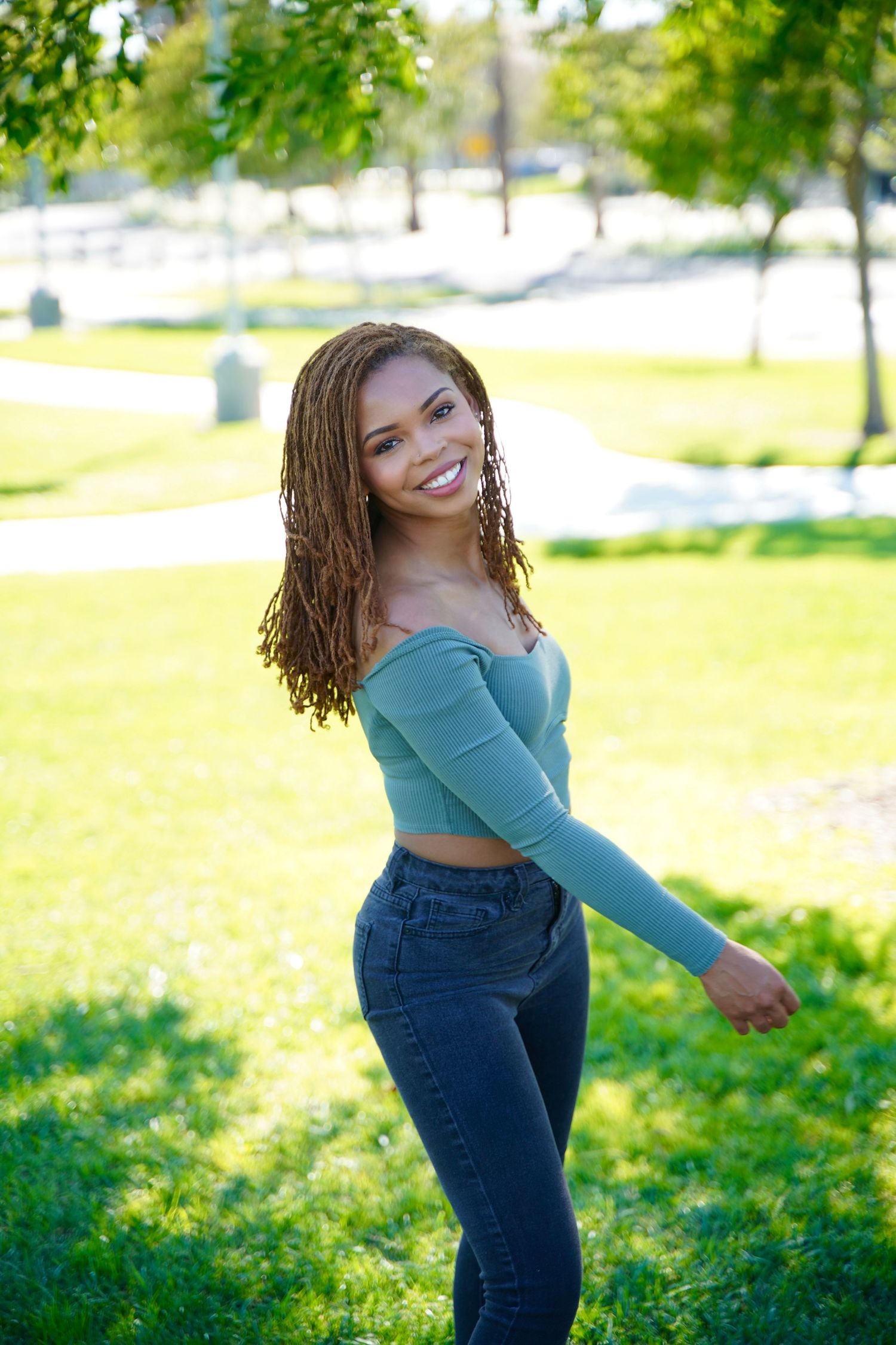 Smiling young Black woman with braids wearing green crop top and jeans posing in sunny park.