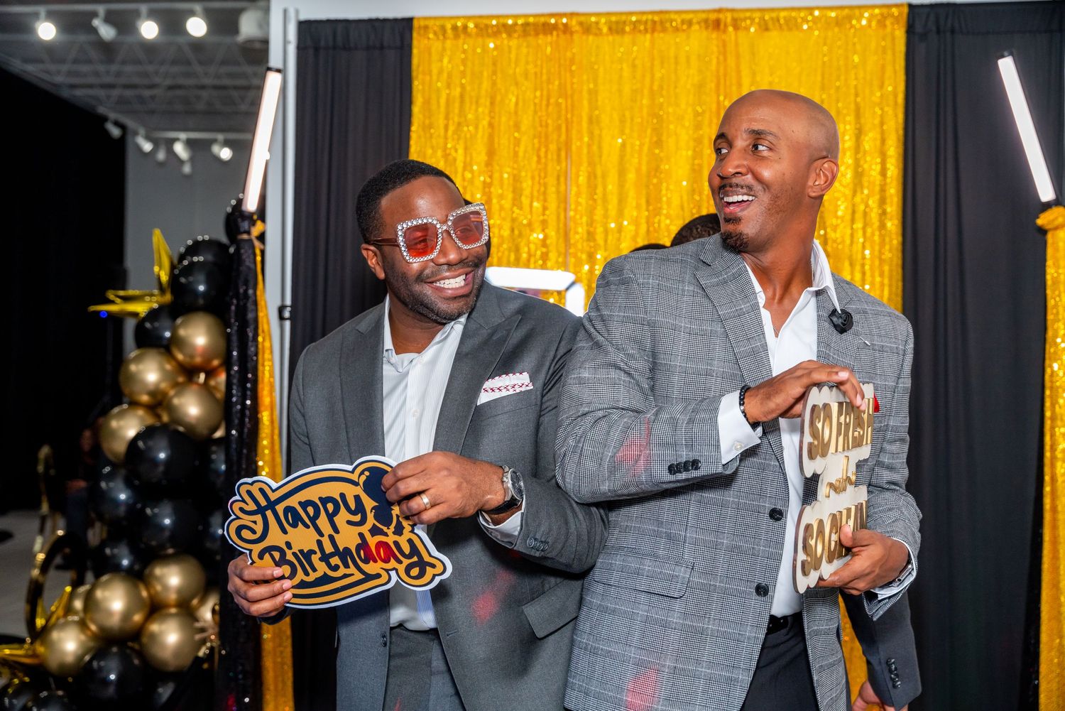 Two men in suits smiling at a birthday party, holding Happy Birthday sign near gold and black balloon decorations.