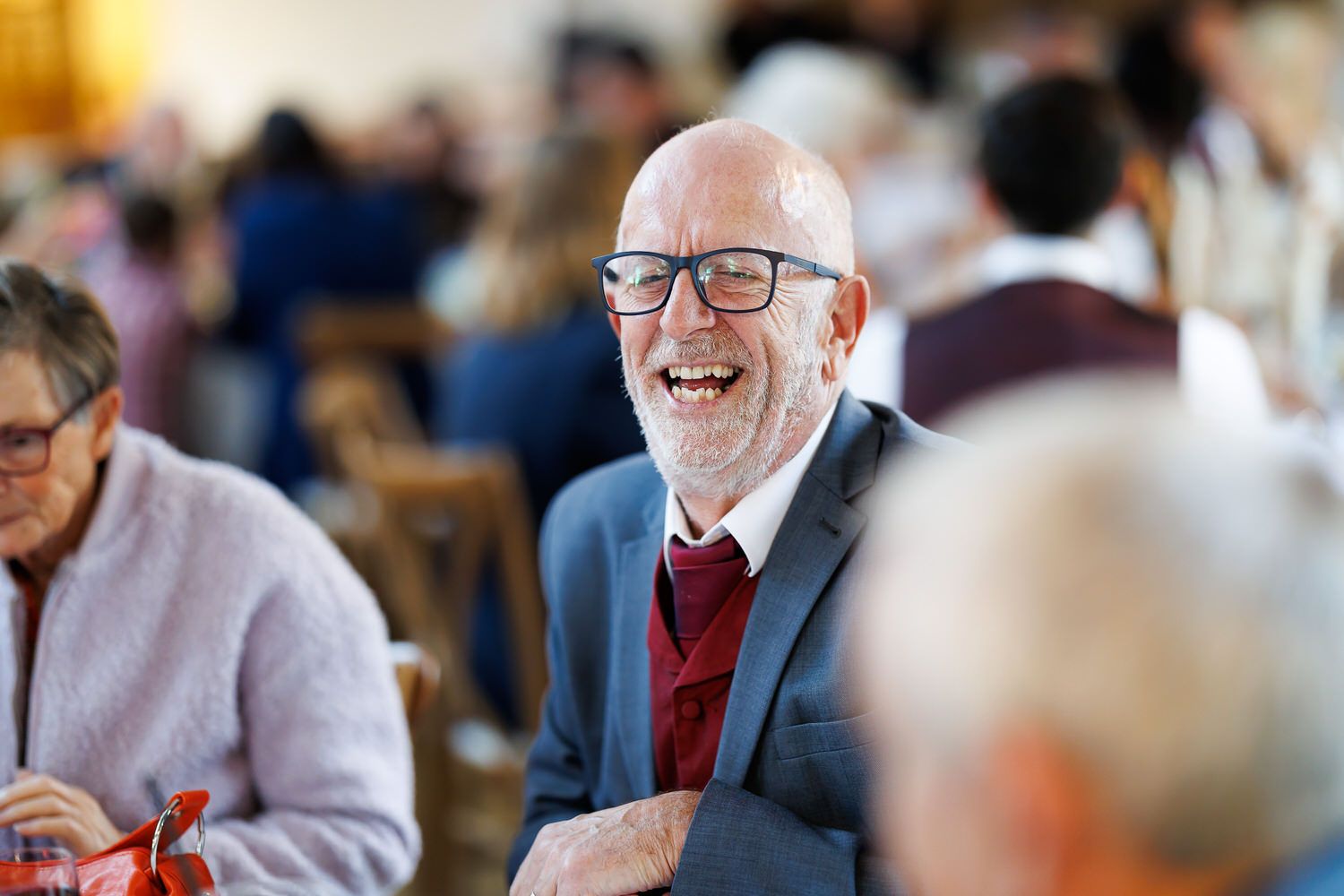 Bald man with glasses laughing joyfully at a social gathering, wearing a gray blazer and red tie.