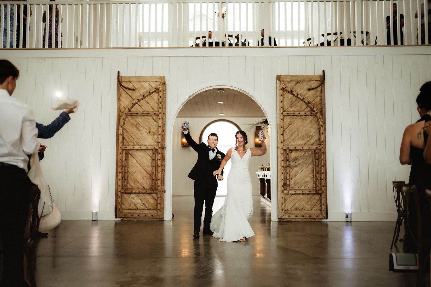 Bride and groom make grand entrance through rustic wooden arch doors at elegant white barn wedding venue.