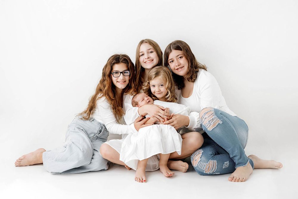 Four sisters sitting together on white background holding a newborn baby in a professional family photo.
