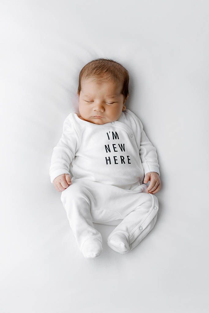 Sleeping newborn baby in white onesie reading I'm New Here on a soft white background.
