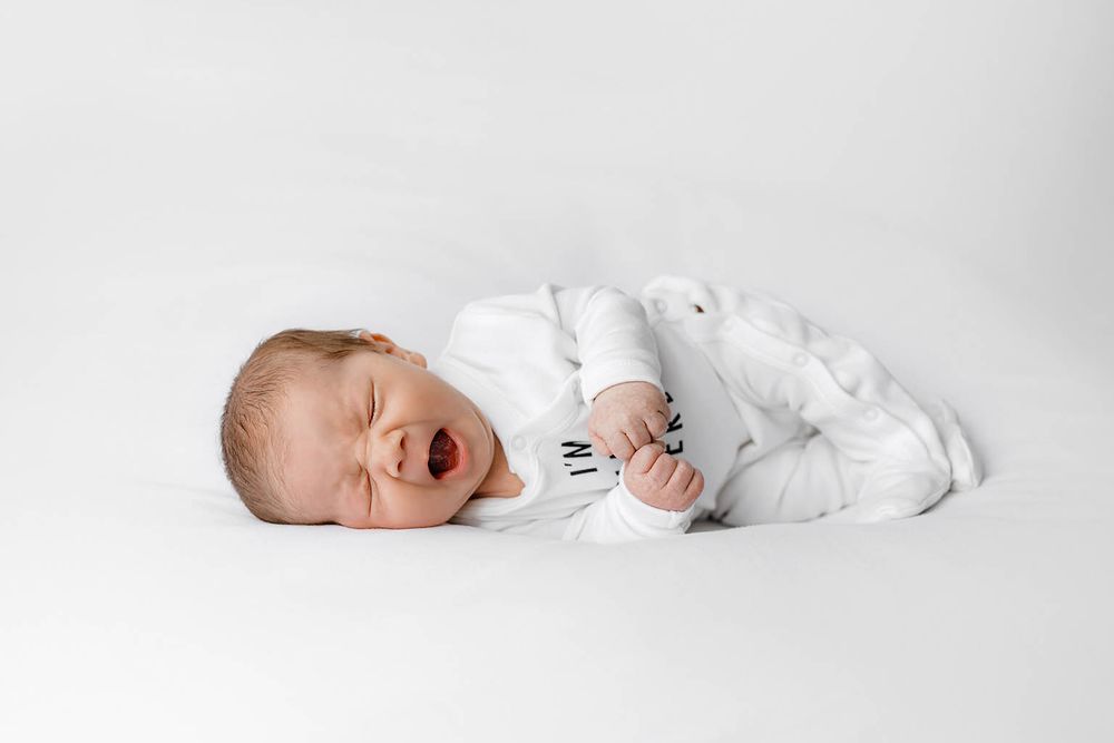 Newborn baby yawning and stretching in a white outfit on a white background.