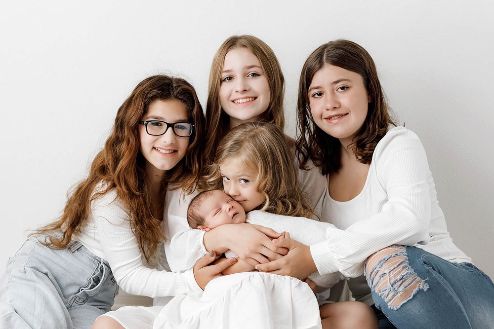 Four sisters in white tops pose together, the oldest three smiling while the youngest holds a sleeping newborn baby.