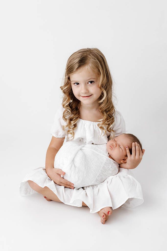 A young girl in a white dress sits cross-legged holding a sleeping newborn baby against a white background.