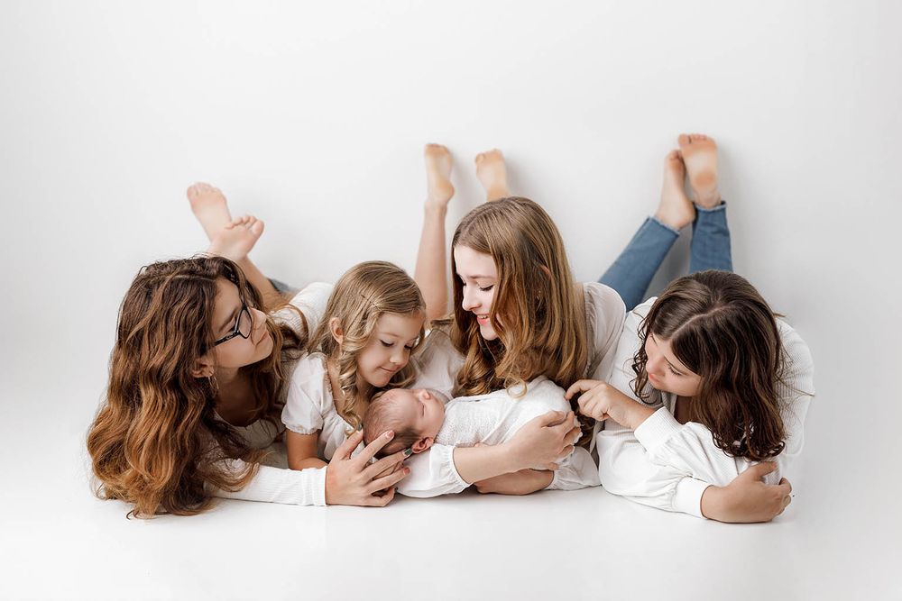 Four girls lying on white floor admiring a newborn baby in a heartwarming family portrait session.