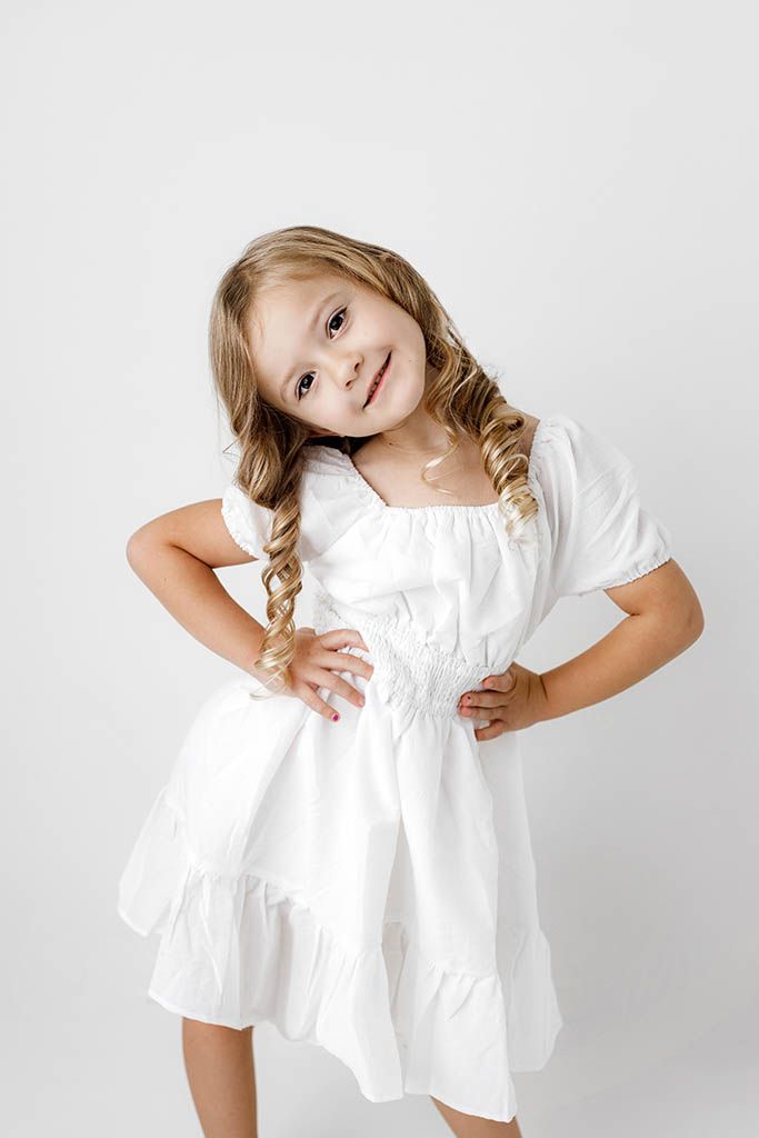 Smiling little girl with braids wearing a white ruffled dress, posing with hands on hips against white background.