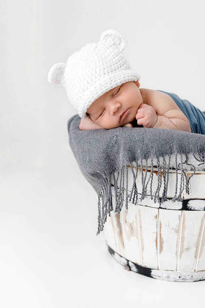 Sleeping newborn baby wearing white knit bear hat resting in rustic white wooden bucket with gray blanket.