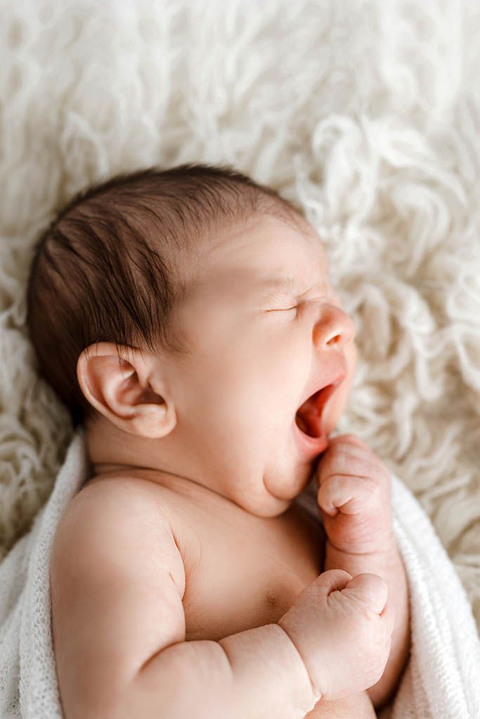 Newborn baby yawning with mouth wide open, lying on a soft white fluffy blanket, eyes closed.
