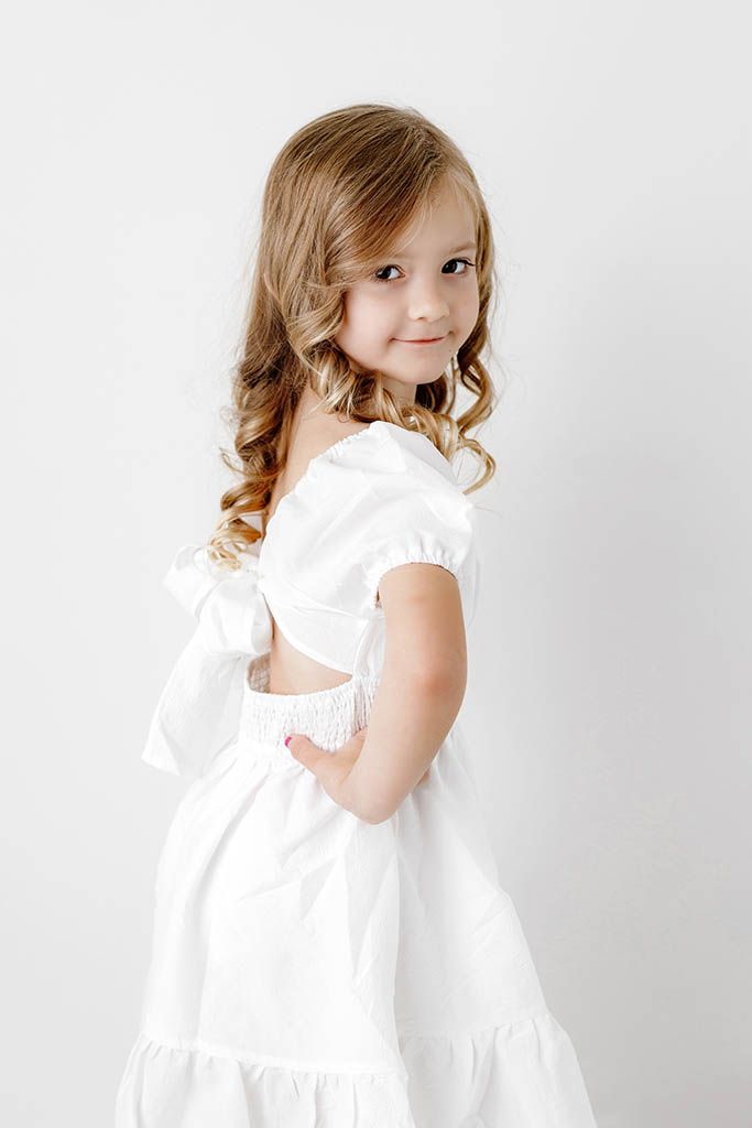 Sweet young girl in a white dress with bow back detail smiling over her shoulder.
