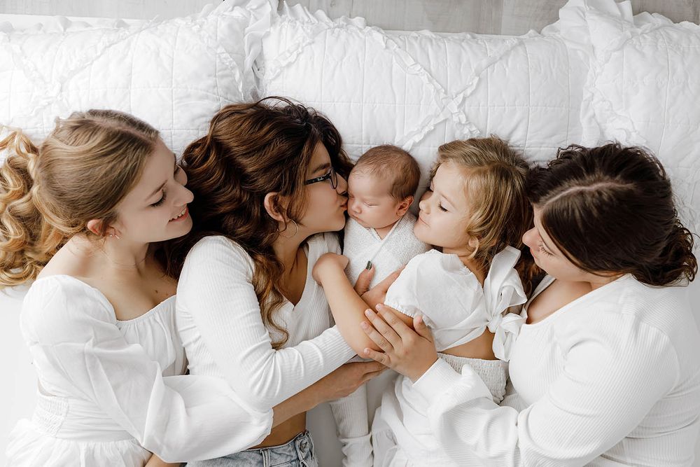 Four women in white clothing cuddle together on a white bed with a newborn baby and toddler.