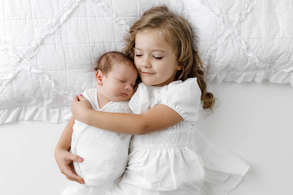 Young girl in white dress tenderly hugging sleeping newborn sibling on white bedding.