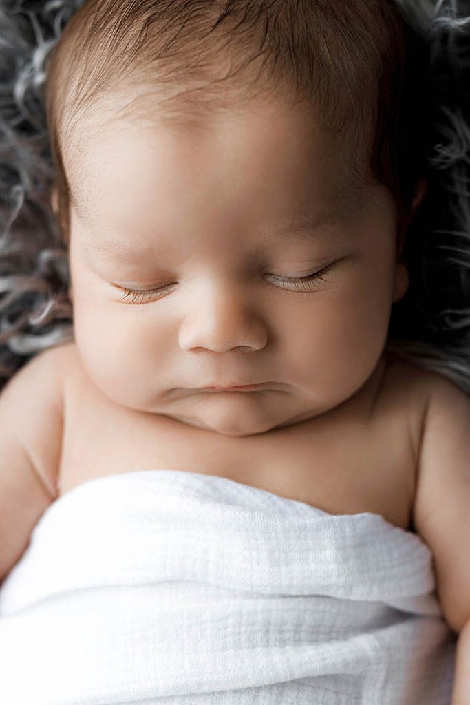 Peaceful newborn baby sleeping wrapped in a white swaddle blanket on a soft gray fluffy surface.