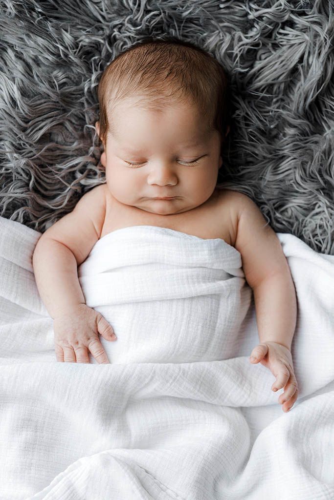 Peaceful newborn baby sleeping wrapped in white muslin cloth on gray fluffy blanket.