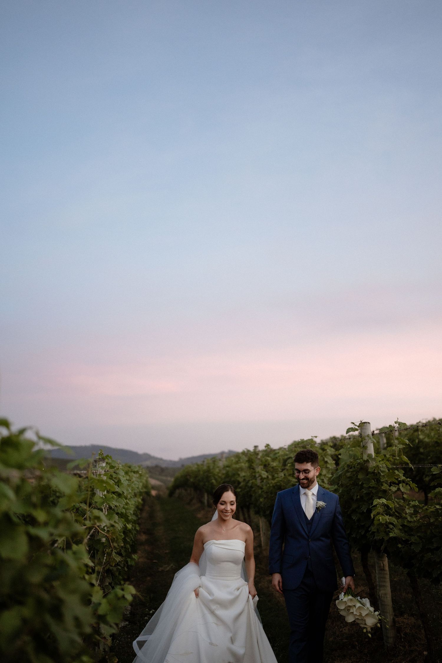 A bride and groom walk hand in hand through a lush vineyard at dusk, with soft pink skies and rolling hills behind them.