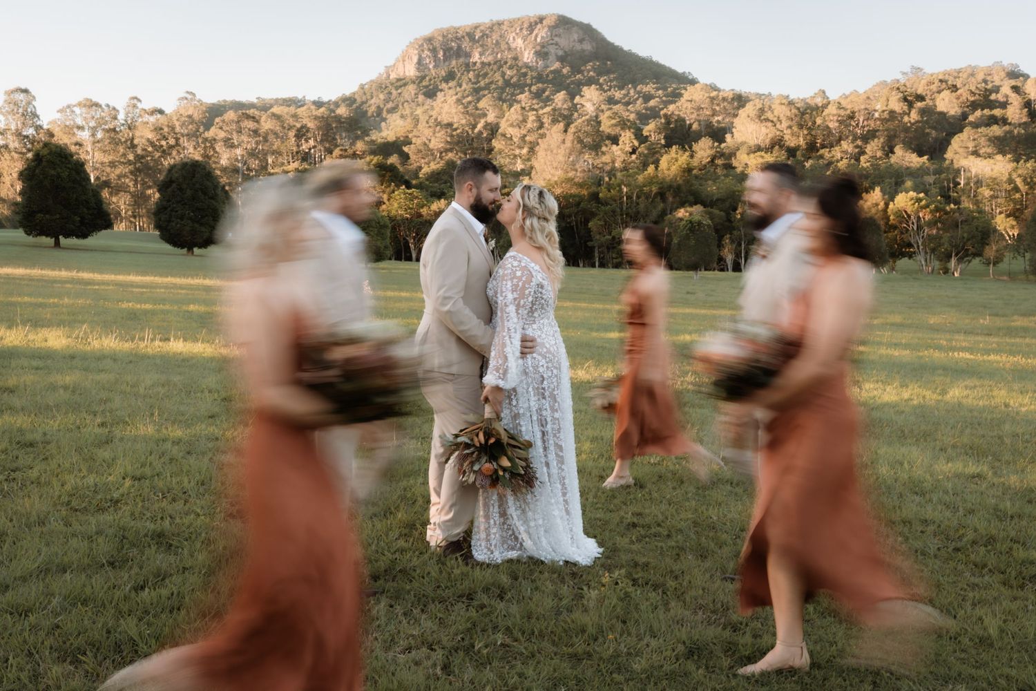 Bride and groom share a romantic moment in a lush green field with blurred bridesmaids in rust dresses passing by.