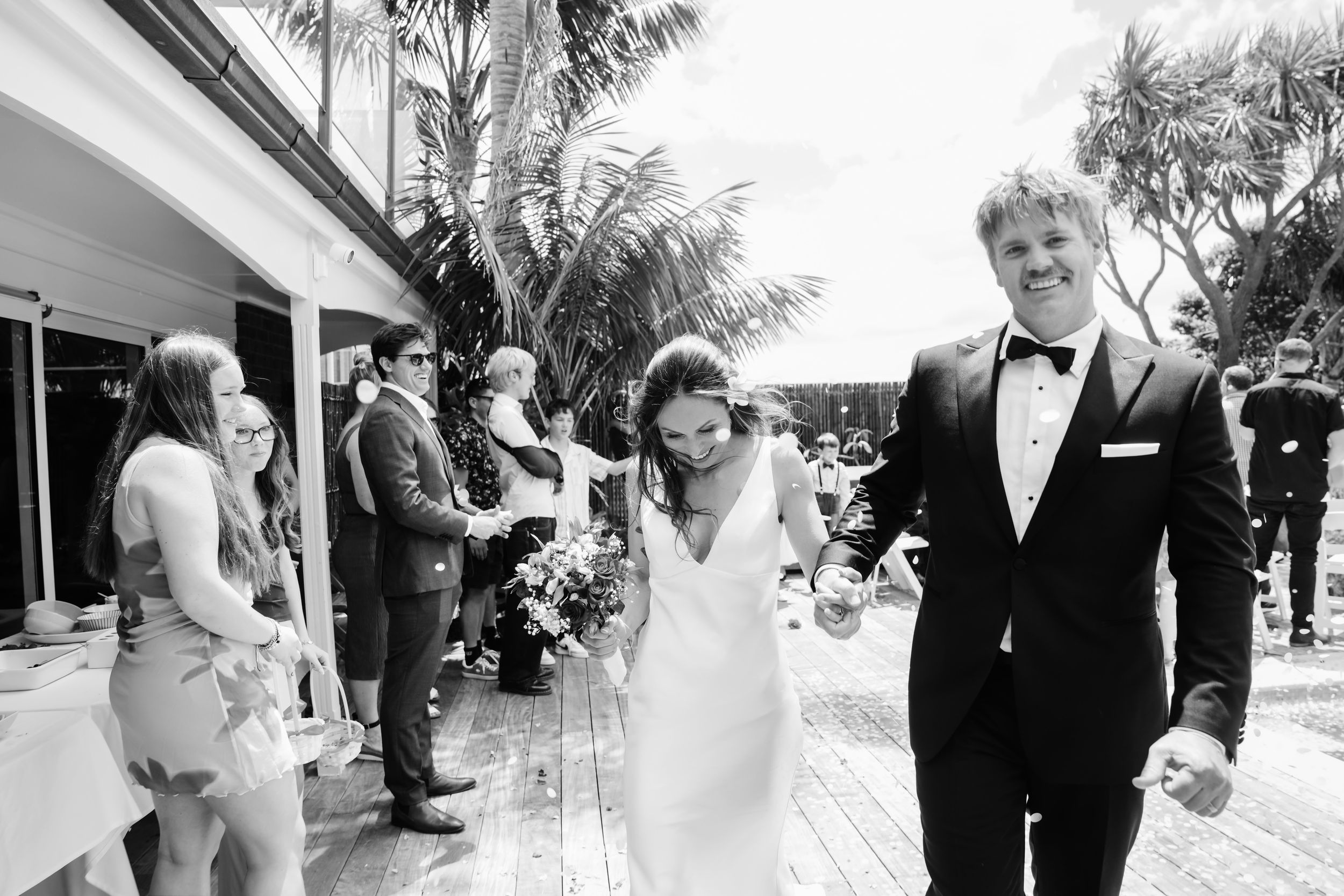 Black and white photo of newlywed couple making grand exit through cheering guests at outdoor venue.
