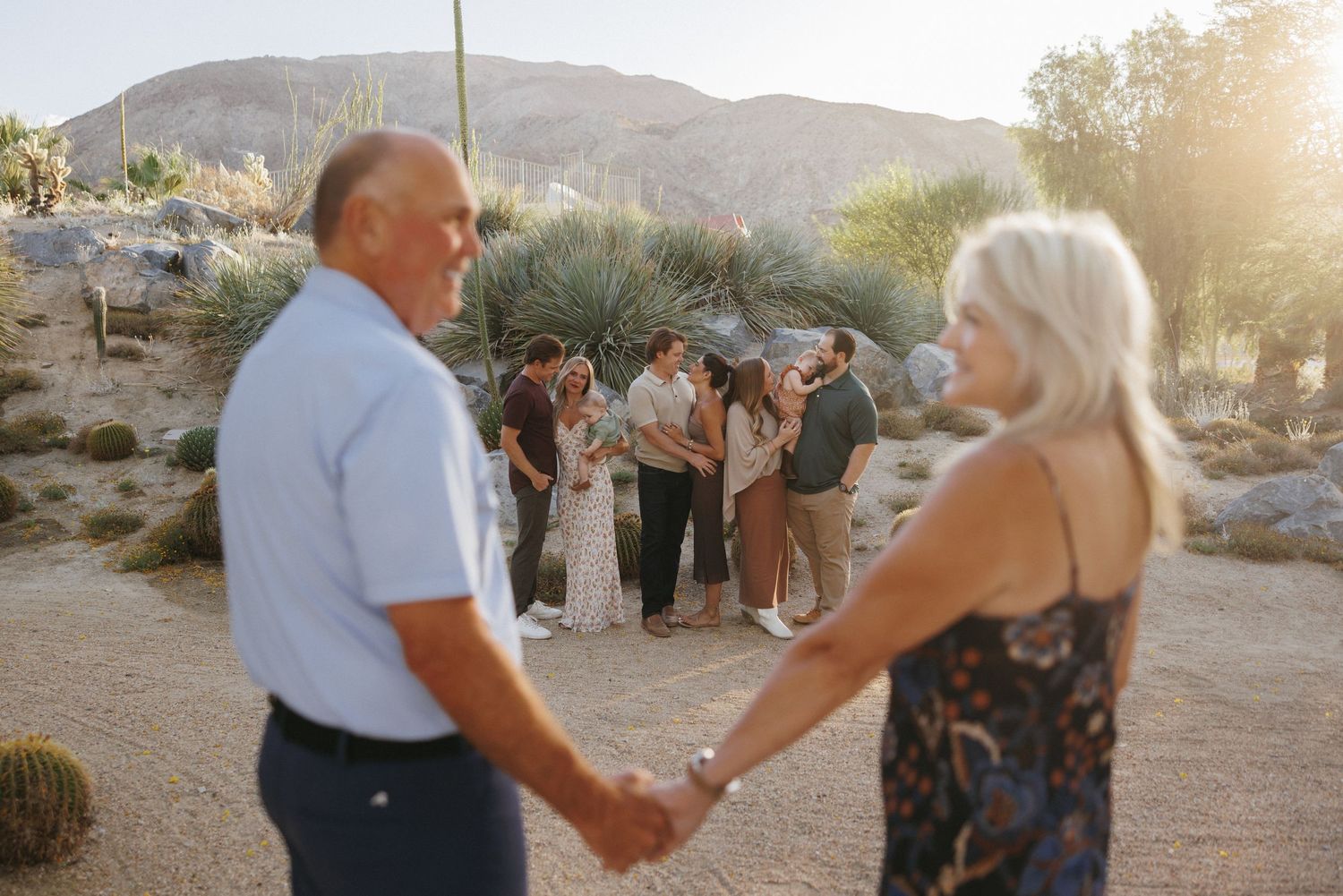 Family of four walks hand-in-hand along a sunny resort path lined with palm trees and blooming bougainvillea.