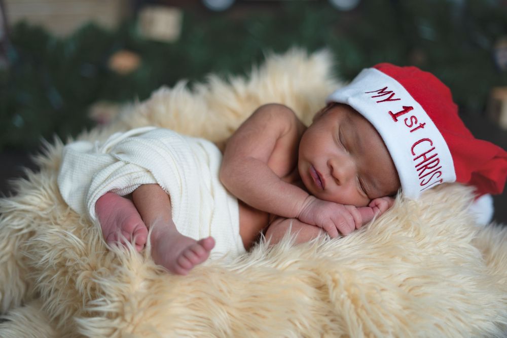 Newborn baby sleeping in a fur basket wearing a red My First Christmas Santa hat holiday photo.