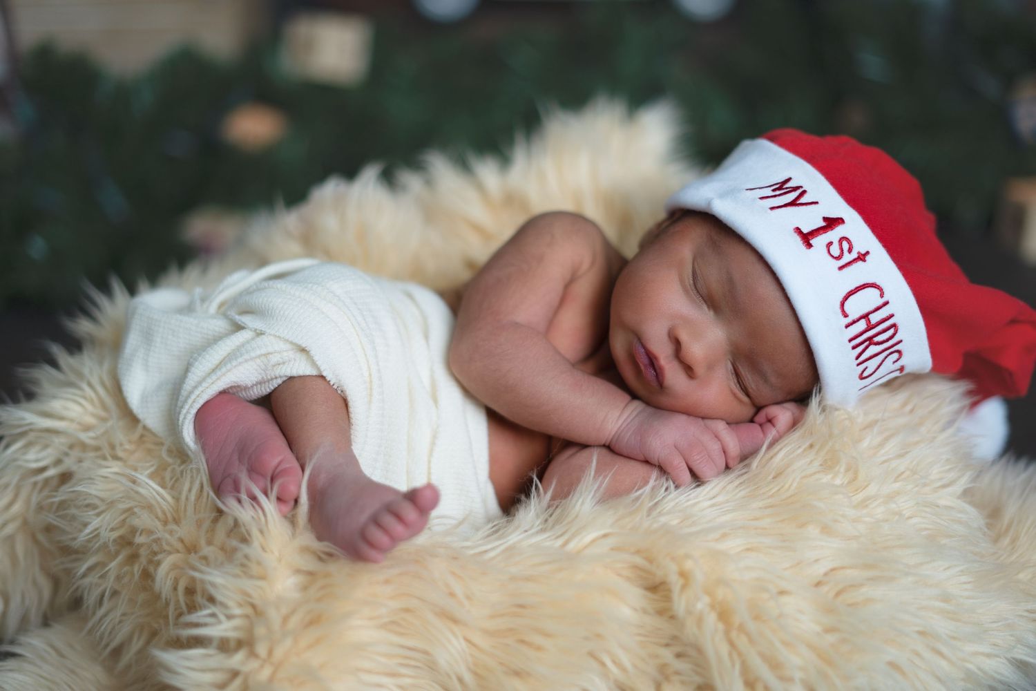 Newborn baby sleeping in a fur basket wearing a red My First Christmas Santa hat holiday photo.