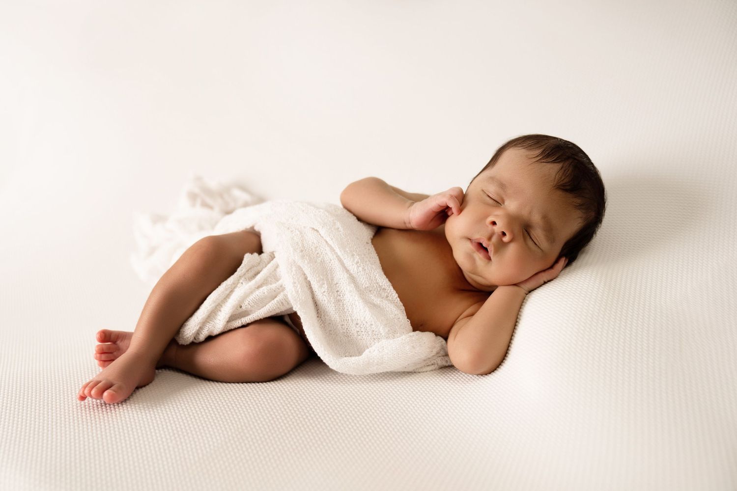 Peaceful newborn baby sleeping curled up on white backdrop, wrapped in soft white cloth.