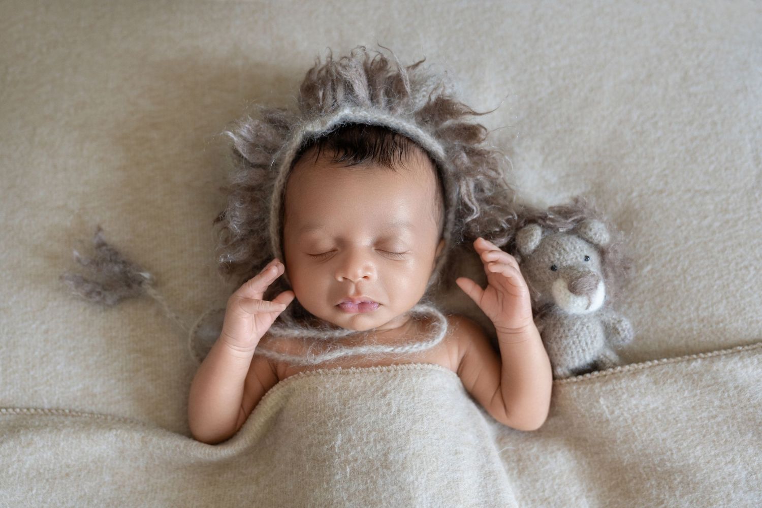 Sleeping newborn baby wearing a lion bonnet hat, wrapped in a cream blanket with a small lion stuffed animal toy.