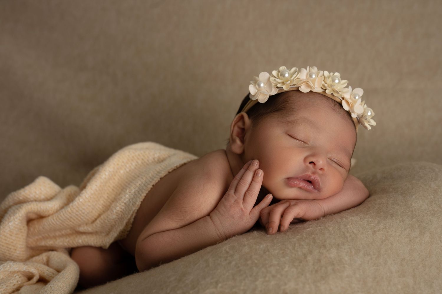 Sleeping newborn baby girl wearing a delicate floral headband, posed on a soft beige blanket in a studio setting.
