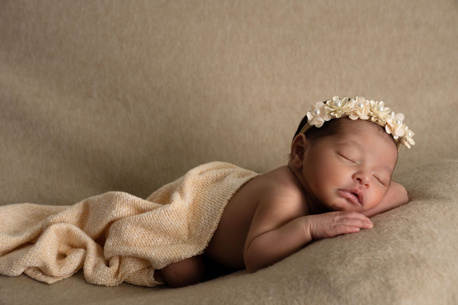 Sleeping newborn baby with floral headband wrapped in beige knit blanket on tan background.