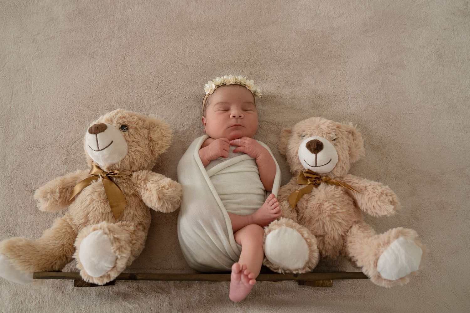 Newborn baby wrapped in white swaddle with floral headband sleeping between two teddy bears on wooden tray.