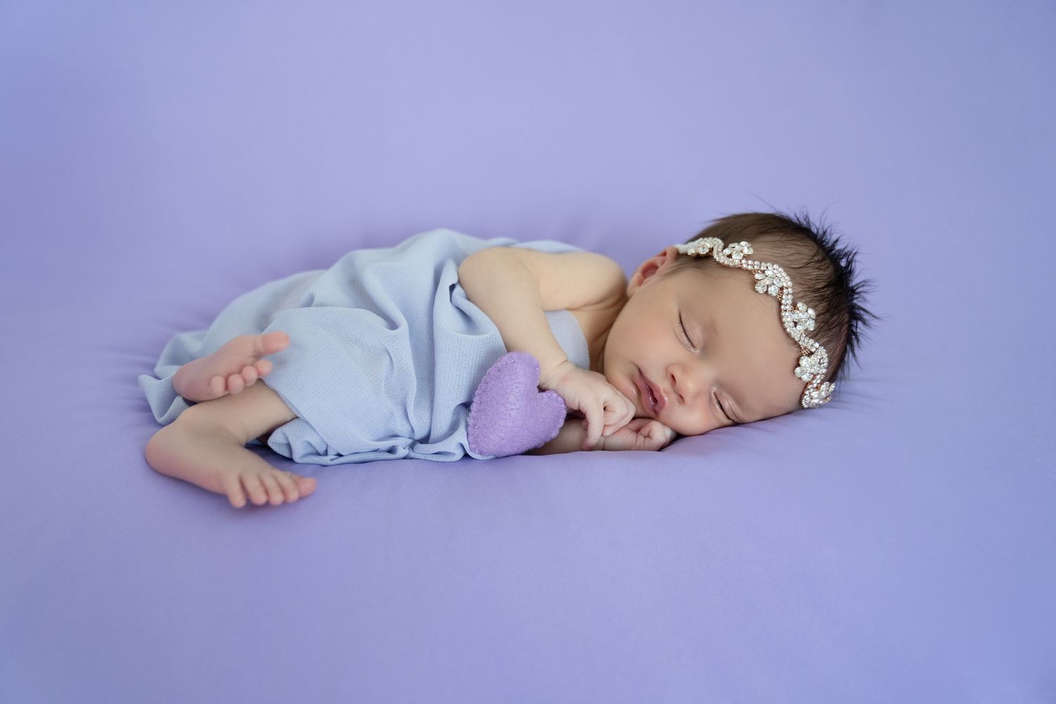 Sleeping newborn baby girl wrapped in lavender cloth, wearing floral headband, holding purple heart on purple background.