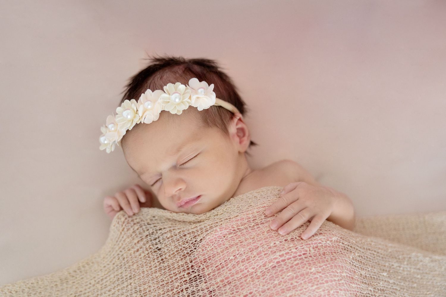 Sleeping newborn baby girl wearing a delicate white floral headband, wrapped in a soft pink knit blanket.