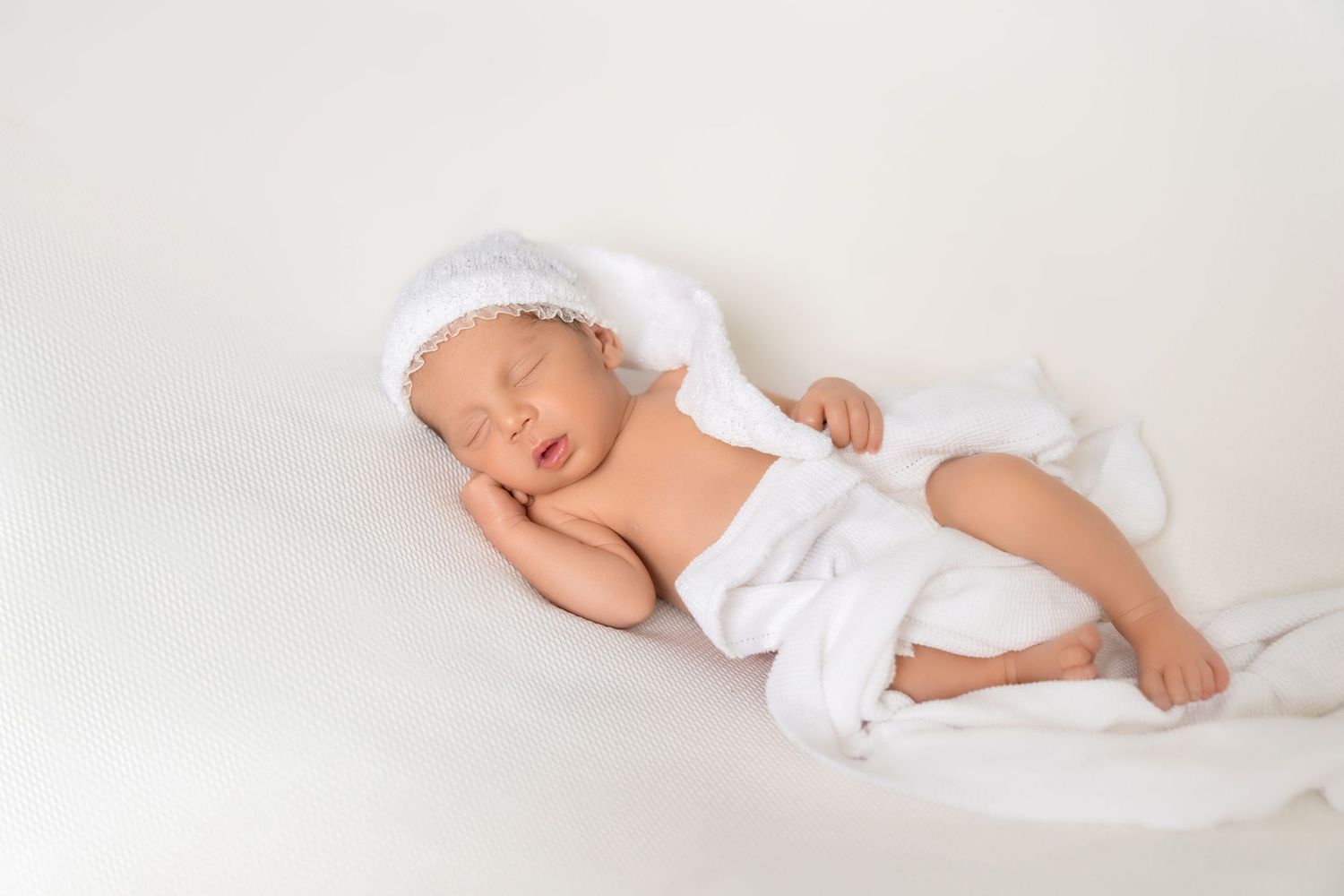 Peaceful newborn baby sleeping in white knit hat and wrap on soft white background.