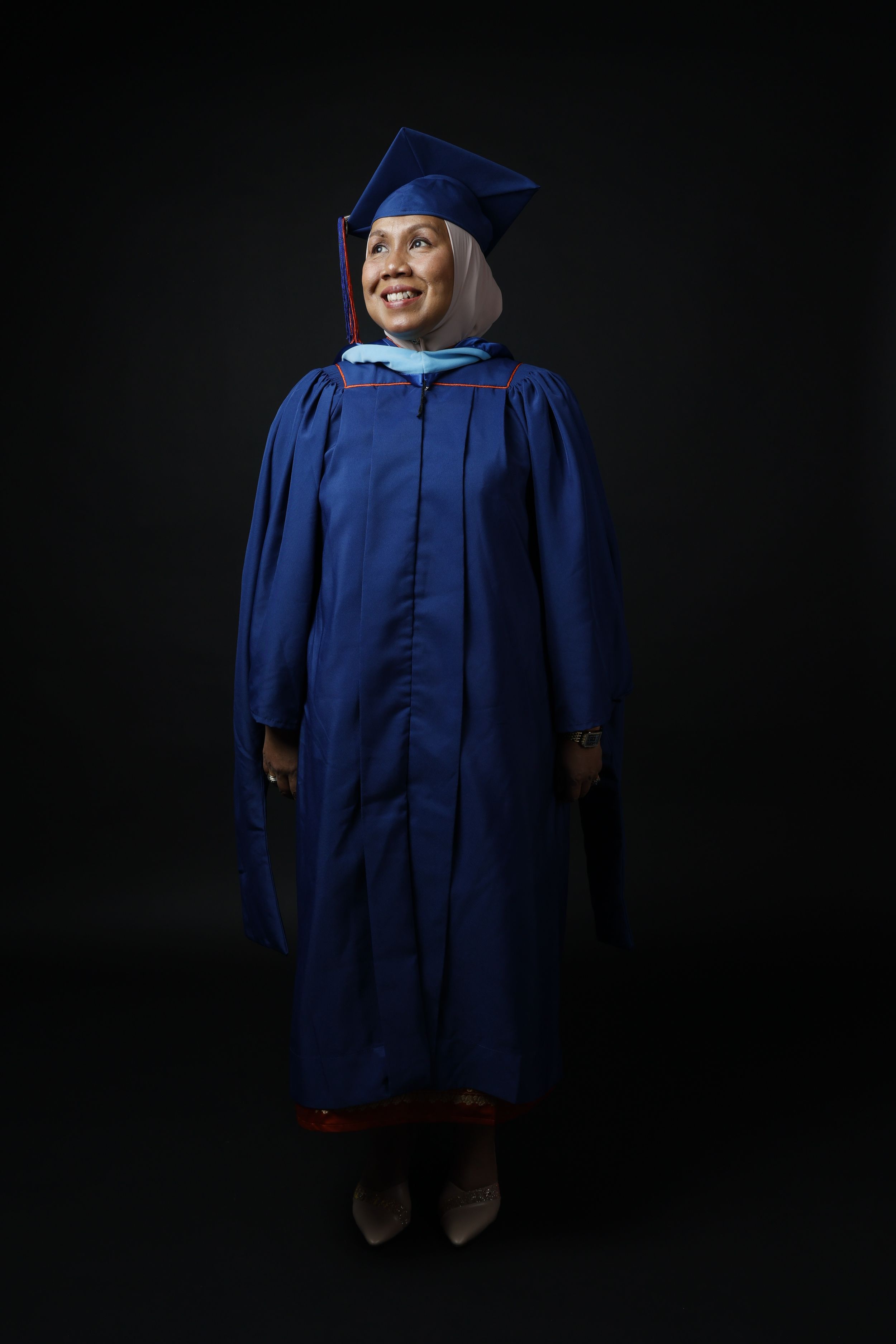 Woman in blue graduation gown and cap smiling, wearing hijab, posing against dark background.