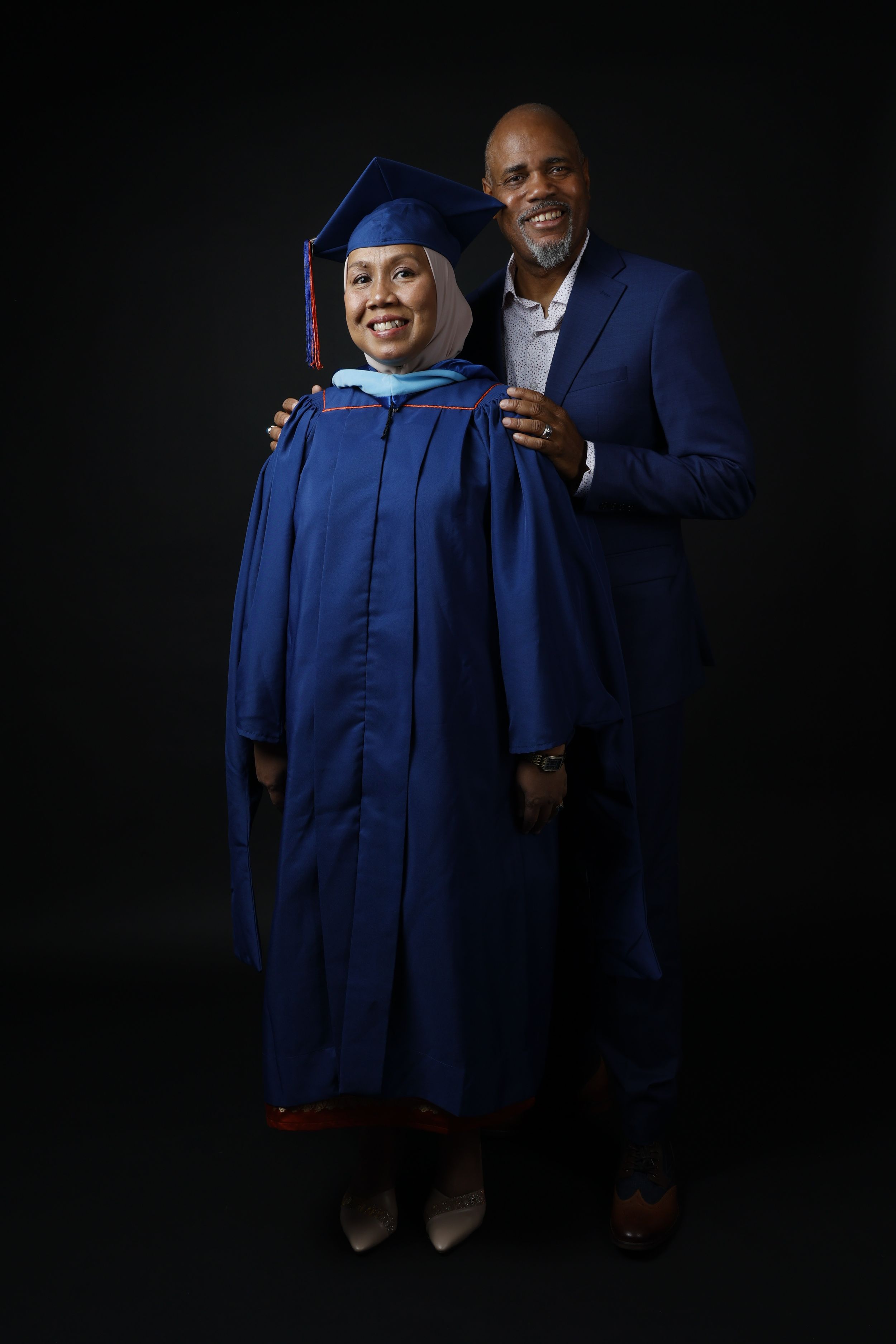 Woman in blue graduation cap and gown poses with man in blue suit against dark background.