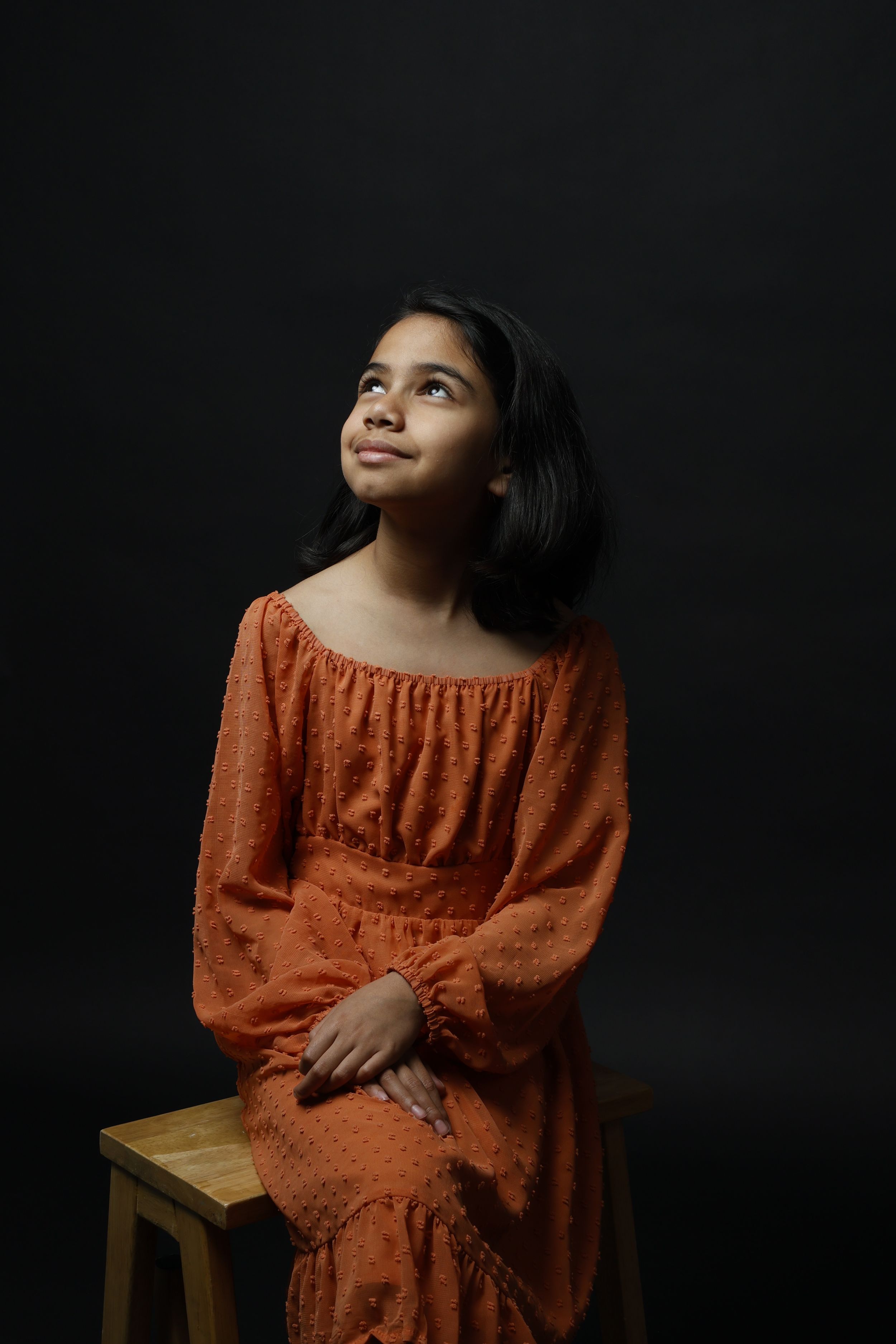 Young girl in orange dress sitting on wooden stool, smiling and looking up against dark background.