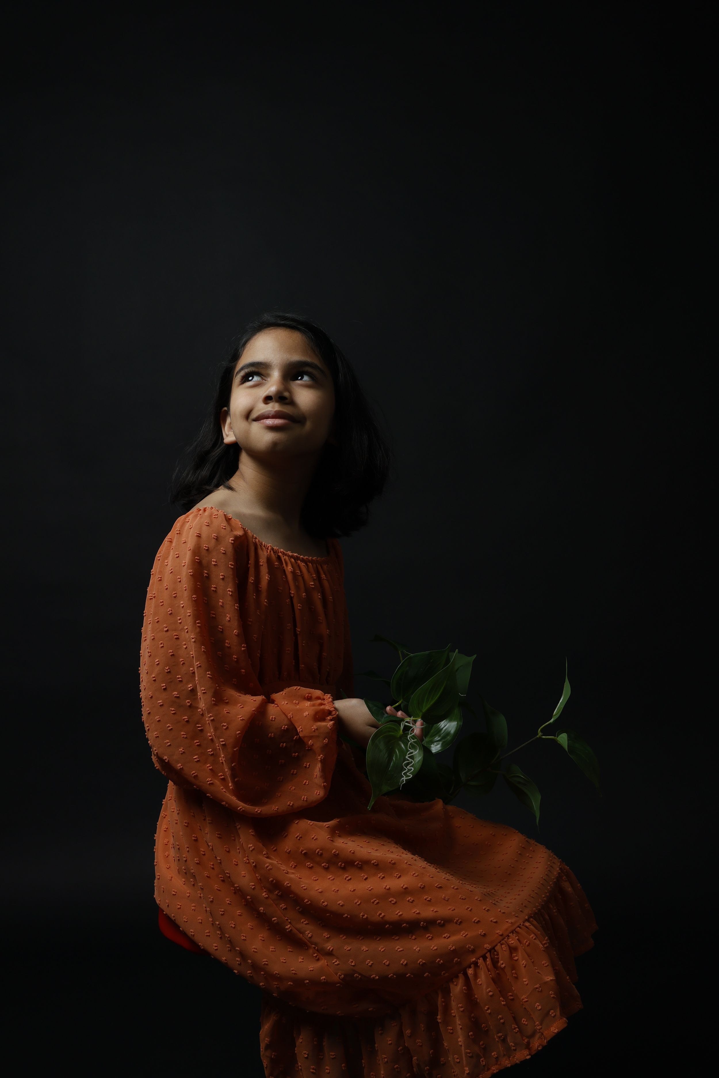 Young girl in orange dress holding green flowers, gazing upward against dark studio background.