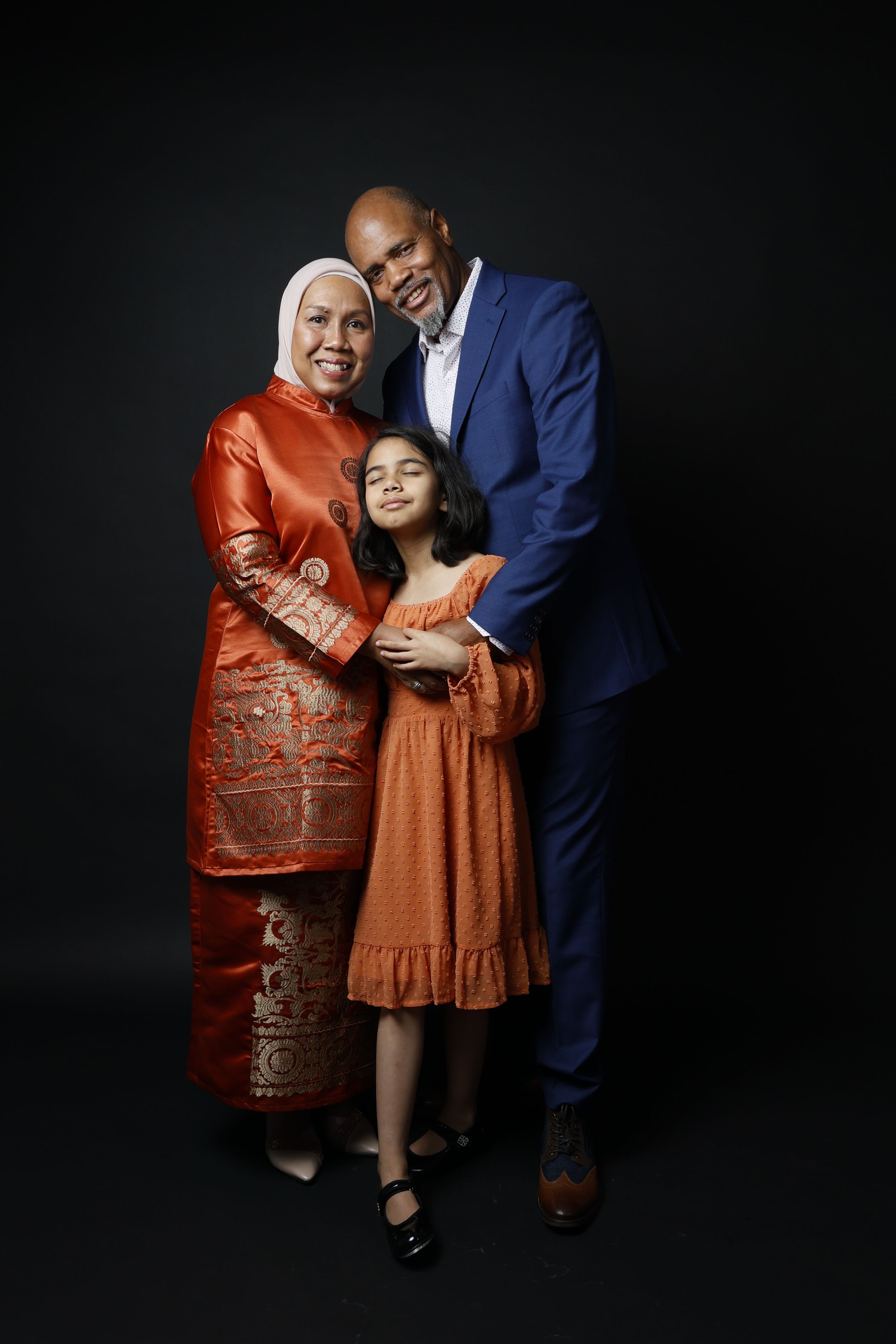 A family of three poses for a studio portrait, with parents in elegant attire embracing their young daughter against a dark background.