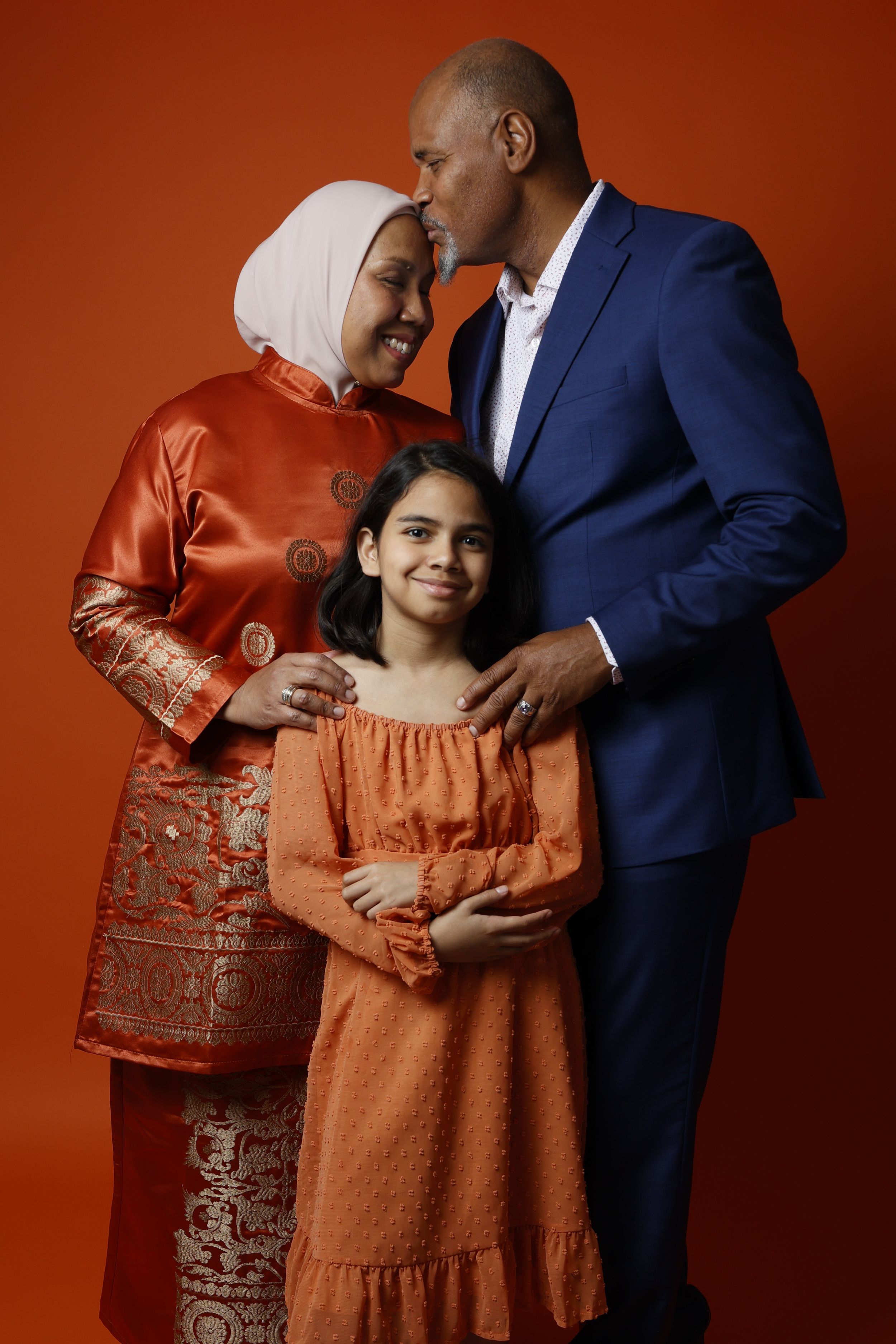 A family of three in orange and blue attire poses warmly together against a vibrant orange background.