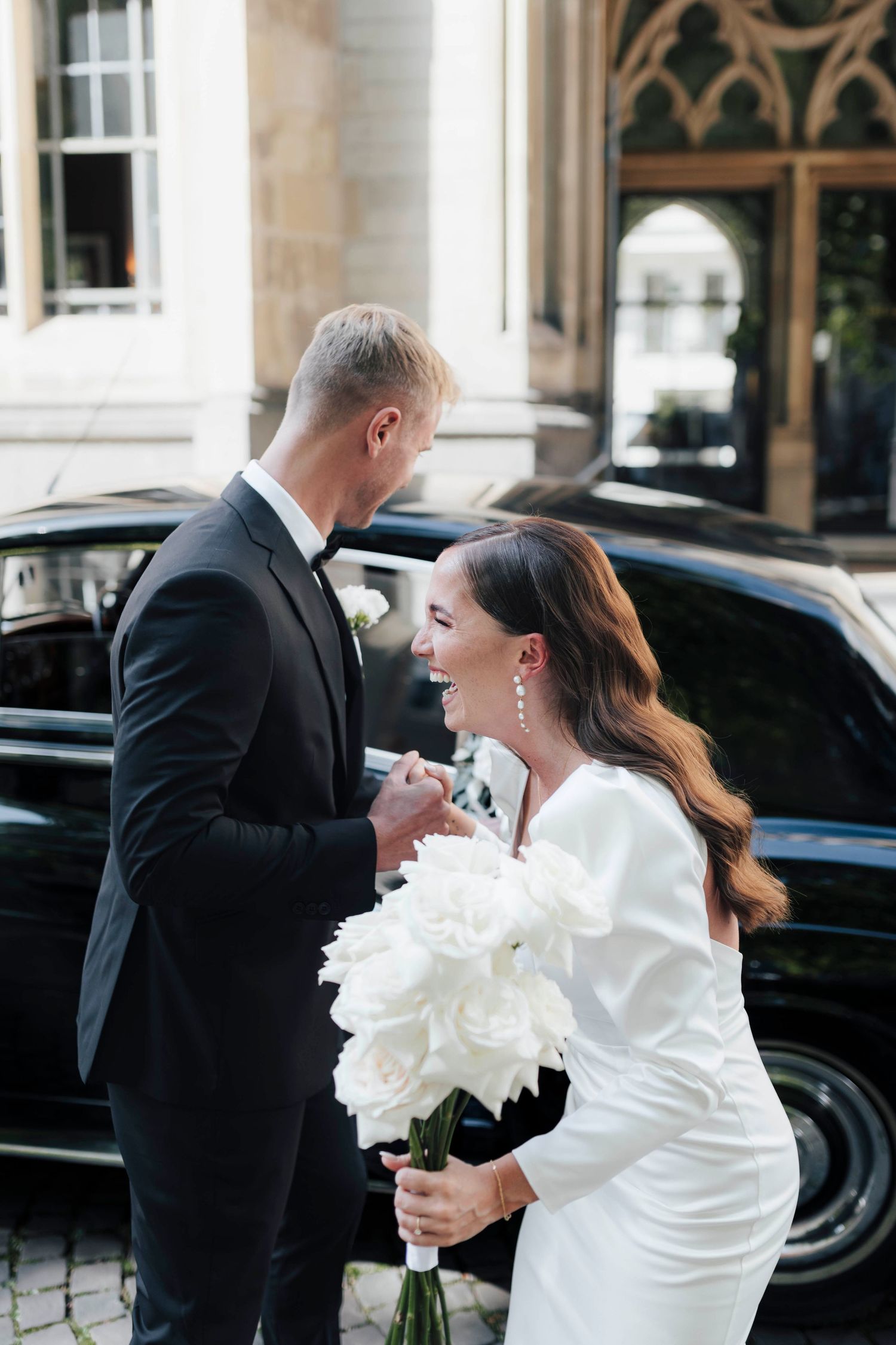 Bride in white gown holding white peony bouquet laughs with groom in black tuxedo outside elegant building.