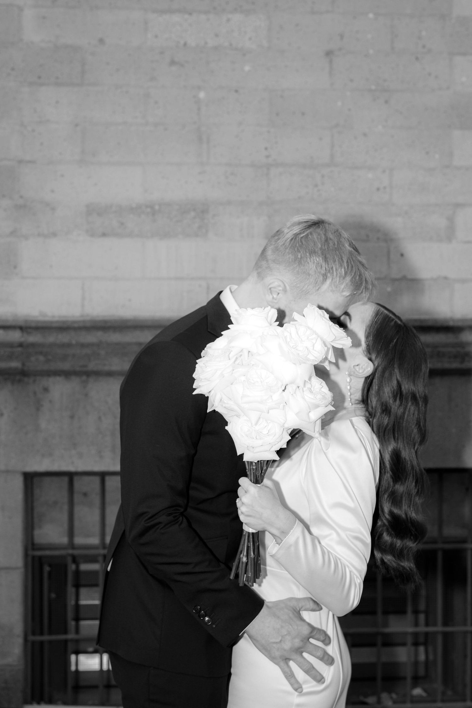 Black and white photo of a couple sharing a kiss, bride holding white floral bouquet in elegant wedding attire.