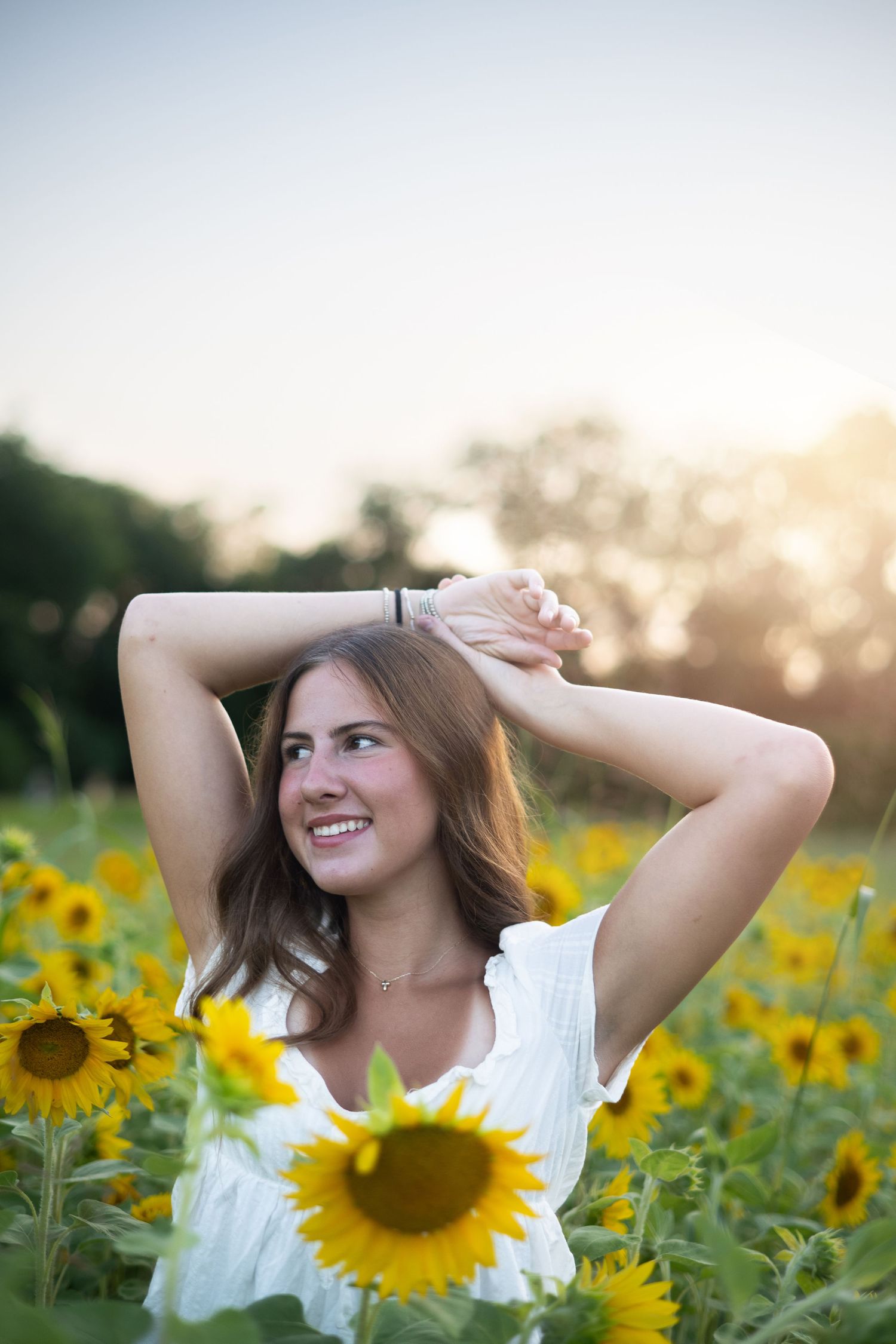 Young woman in white dress smiling in a sunflower field at golden hour with hands raised above her head.