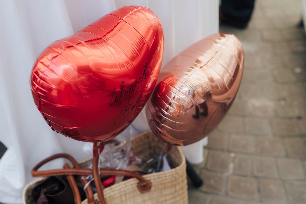 Two heart-shaped foil balloons, red and rose gold, displayed in a wicker basket near a white curtain.
